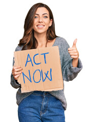 Young brunette woman holding act now banner smiling happy and positive, thumb up doing excellent...