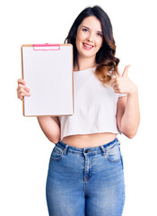 Beautiful young brunette woman holding clipboard with blank space smiling happy and positive, thumb up doing excellent and approval sign