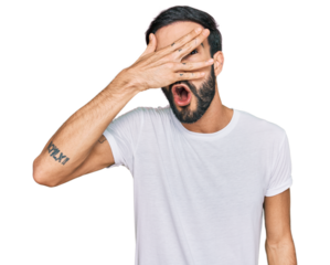 Young hispanic man wearing casual white t shirt peeking in shock covering face and eyes with hand, looking through fingers with embarrassed expression.