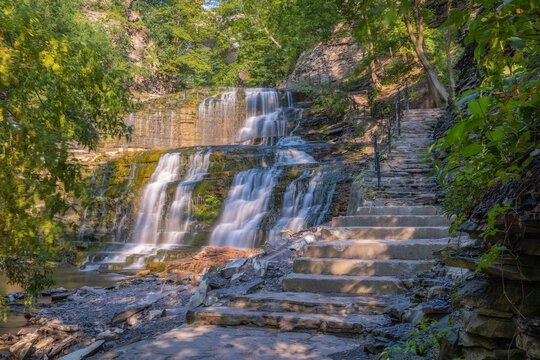 Stairs leading up a beautiful cascading waterfall surrounded by lush greenery. Cascadilla Gorge in Ithaca New York- Finger Lakes Region
