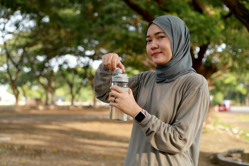 Young veiled asian muslim woman in posing resting after training hold bottle of water, looking at the camera. Outdoor sport concept