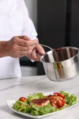 Professional chef holding saucepan with sauce near delicious dish at white marble table in kitchen, closeup