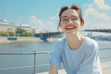 Fototapeta premium Young woman smiling joyfully on a riverside walk with a cityscape backdrop on a sunny day.