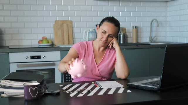 A young woman shakes coins out of a pink piggy bank onto a kitchen table, scattering money. Concept of managing personal finances and budgeting