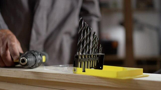 Close-up of experienced carpenter who chooses a drill for drilling a wooden plank working in carpentry shop