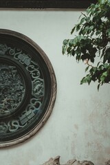 A round window with intricate carvings depicting figures on horseback, set within a wooden frame, against a white wall. Green leaves of a tree branch partially obscure the right side of the image.