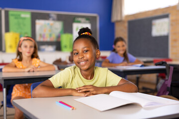 Portrait of cute smiling african american schoolgirl with book on desk studying in classroom