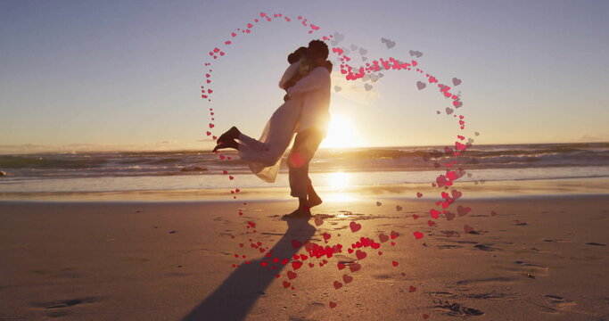 Image of hearts over happy diverse just married couple dancing on beach by sea