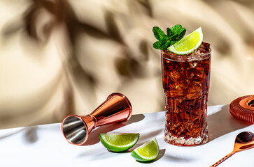 Rum cola cocktail long drink with ice, mint and lime in glass. Beige background