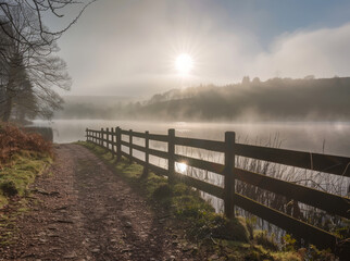 Naklejka premium A misty morning in the lake district, a wooden fence along an amber woodland trail leading to water and a distant sun breaking through the fog... 