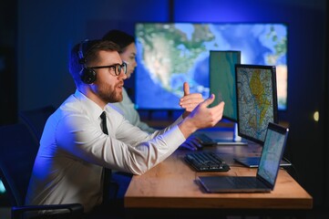 In Monitoring Room Technical Support Specialist Speaks into Headset. His Colleagues are Working in...