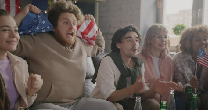 Multiethnic group of young friends fans watching sports waving American flags cheering enjoying game at home. Patriots and entertainment concept.