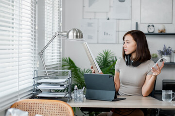 A young woman multitasking in a home office, using a tablet and smartphone, surrounded by modern office supplies and natural light.