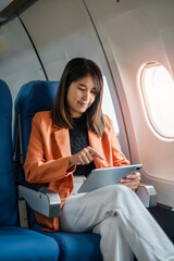 A businesswoman in a bright orange blazer uses a tablet while seated on an airplane, highlighting in-flight productivity and modern travel.