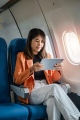 A young woman in a bright orange jacket uses a tablet while seated in an airplane, enjoying her flight.