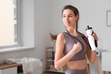 Beautiful young happy sporty woman with bottle of water and fitness expander at home