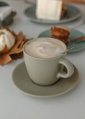 Cappuccino with whipped thick milk foam in a ceramic cup on a saucer with a spoon stands on a table in a cafe. Autumn decor with fall leaves and dessert, macaroon on the plate are next to the coffee