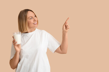 Fototapeta premium Beautiful young woman with glass of fresh milk pointing at something on beige background