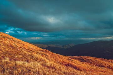 A scenic landscape view of a mountain range with a dramatic sky. Foreground of rolling hills with dry grass and a distant mountain range covered in clouds. The sky is a vibrant blue with heavy
