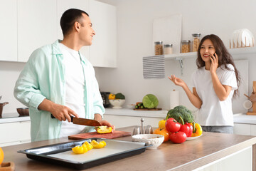 Young man cutting bell pepper with his wife talking by mobile phone in kitchen