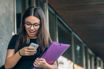 Girl university student user holding cell phone using mobile applications tech on smartphone looking at cellphone, chatting, texting, checking social media, educational apps standing outside campus.