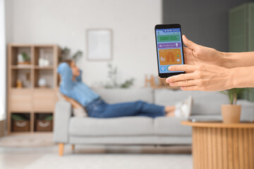 Woman holding mobile phone with smart home security system control panel in room, closeup
