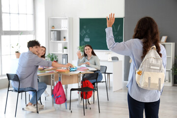 Female teenage student with backpack waving hand to her classmates in classroom, back view