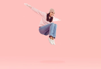 Funny young woman in winter mittens and hat laughing and having fun jumping high. Cheerful Caucasian woman in casual clothes levitating with bent legs and outstretched arms isolated on pink background
