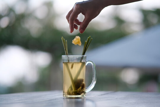 A woman's hand drops a piece of rock sugar into a glass containing hot ginger tea