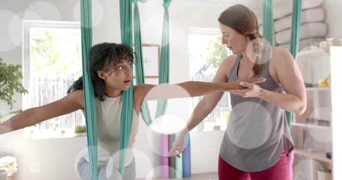 Image of light spots over diverse female instructor and teenage girl practicing aerial yoga - Powered by Adobe