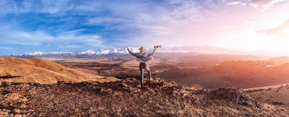 Back view young man traveler with camera on against amazing landscape with mountains Altai, sunset...