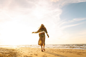 Stylish woman walks at sunset along the beach of the sea or ocean on at sunset. Cold sunny and windy day. Travel, weekend, relax and lifestyle concept.