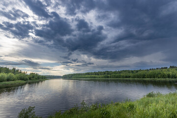 A scenic view of a tranquil river under a dramatic summer sky, with thick, dark clouds gathering overhead and a line of lush green trees along the banks.