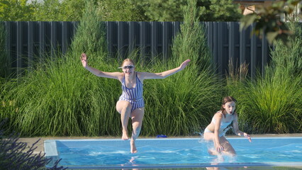 Two teenage girls in swimsuits joyfully jumping into a backyard pool on a sunny day.