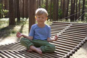 Blonde boy sitting in lotus position and meditating on a hanging wooden hammock in the forest