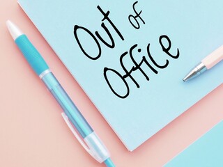 Vibrant composition with a blue paper with the handwritten words out of office on a pink background next to a pair of pens. Simple and minimalistic out-of-office message in pastel colors