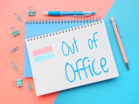 Vibrant composition with a blue paper with the handwritten words out of office on a pink background next to a pair of pens. Colorful out-of-office message in pastel colors