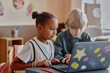Two focused primary school children of diverse ethnic backgrounds using in-class laptop together utilizing online learning tools sitting at table