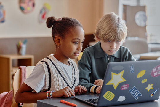 Two focused primary school children of diverse ethnic backgrounds using in-class laptop together utilizing online learning tools sitting at table