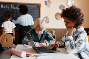 Obraz premium Side view of focused Black girl using laptop in diverse primary school while learning diligently at desk in class, copy space