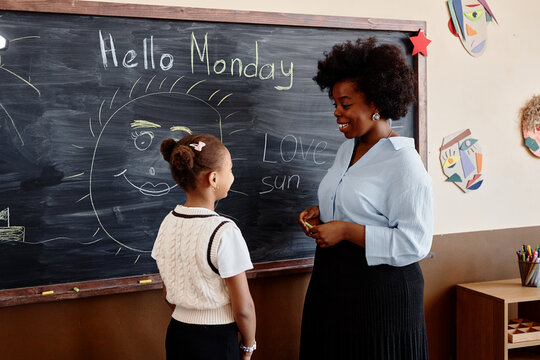 Side view of supportive female teacher of Black ethnicity smiling while listening to little girl student answering at blackboard, copy space