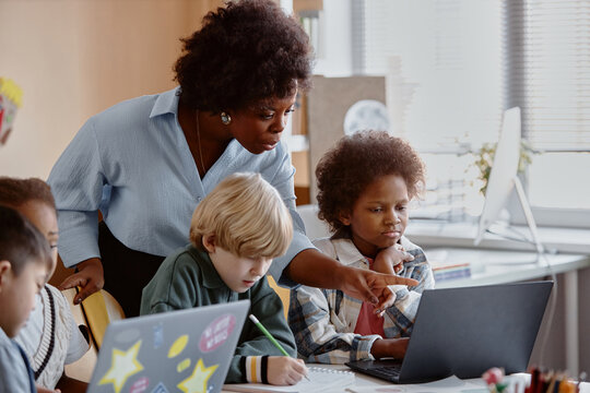 Young female teacher of Black ethnicity explaining new topic to girl student interacting with edtech in diverse group of children at school