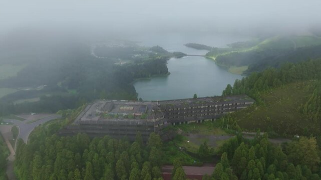 Aerial view of lush landscape with abandoned hotel, serene lake, misty forest, and winding road, Sete Cidades, Azores, Portugal.