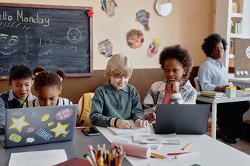 Shot of multiethnic group of children studying in pairs at large desk using laptops in primary school classroom decorated with blackboard and kids crafts