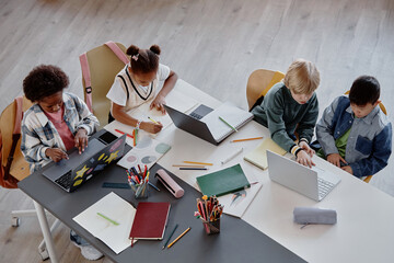 High-angle shot of multiethnic primary students working in pairs writing at large desk while using laptops during class