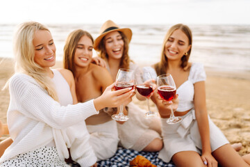 Smiling young women clinking glasses of wine and spend time together. Hen party on the beach. Picnic, vacation, relax, holiday.
