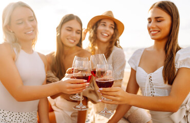 Smiling young women clinking glasses of wine and spend time together. Hen party on the beach. Picnic, vacation, relax, holiday.