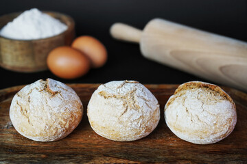 Small fresh breads on a wooden tray next to eggs, a wooden bowl with flour and a rolling pin
