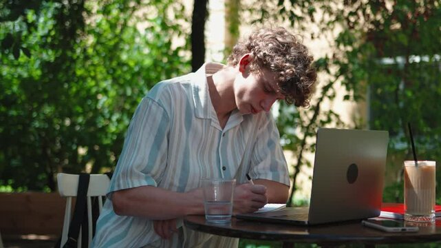 young student male writer writing thinking outdoor at cafe thoughtful hispanic curly hair freelancer working writing with pen, sitting outdoors at cafe with laptop