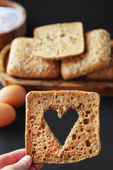 A cut out heart on a slice of bread next to small fresh breads with seeds and sesame on a wooden tray next to eggs, a wooden bowl with flour and a rolling pin